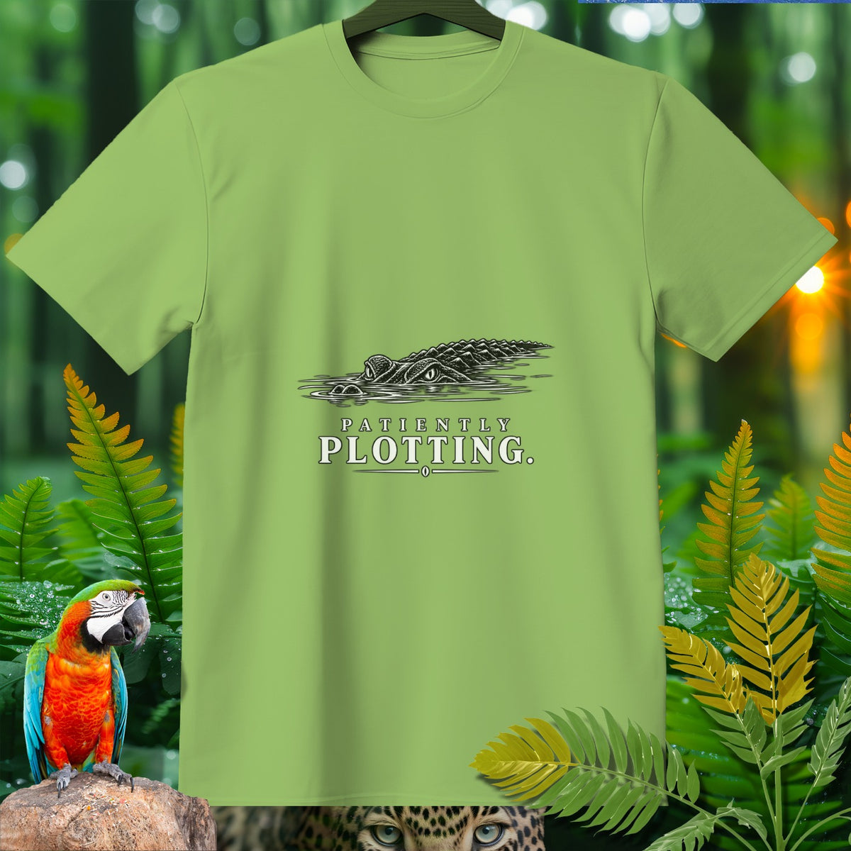 Light green t-shirt with an alligator graphic and text that reads "PATIENTLY PLOTTING."