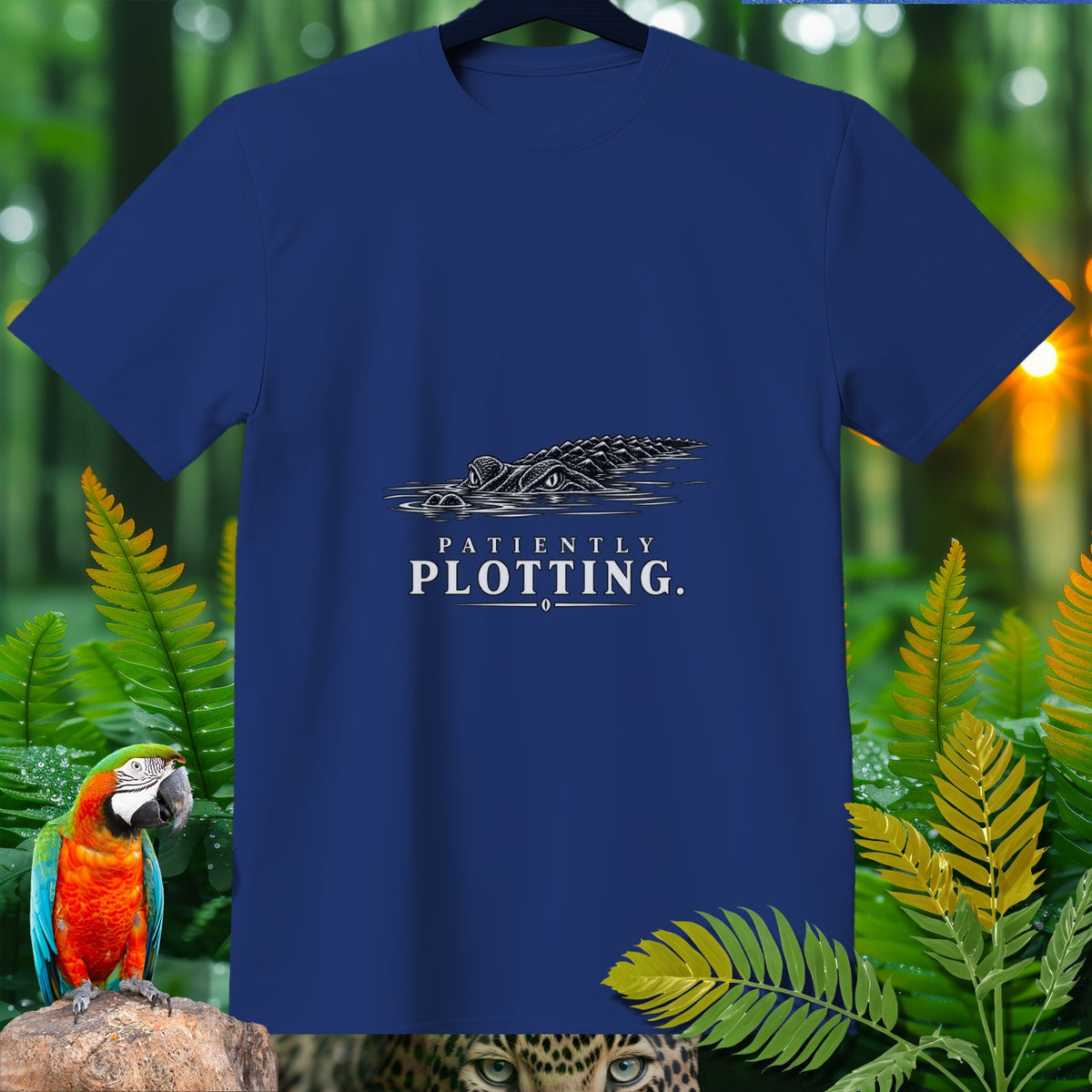 Blue t-shirt with an alligator graphic and the text "PATIENTLY PLOTTING.".